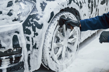 Male worker in uniform washing new modern car that covered with soap. Conception of service