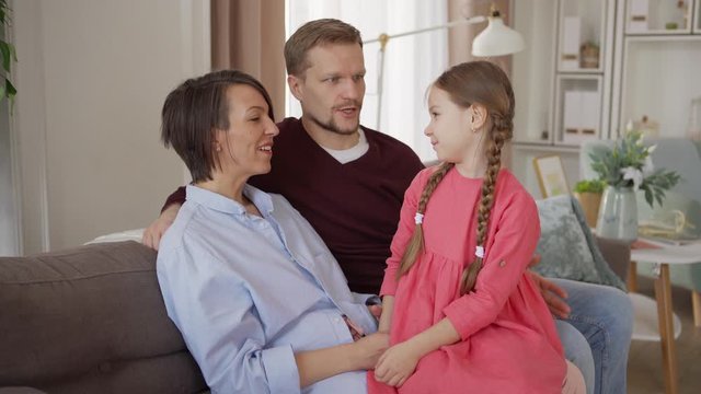 Medium shot of loving middle aged mother and father enjoying talking with cute little daughter sitting on mothers lap on sofa at home