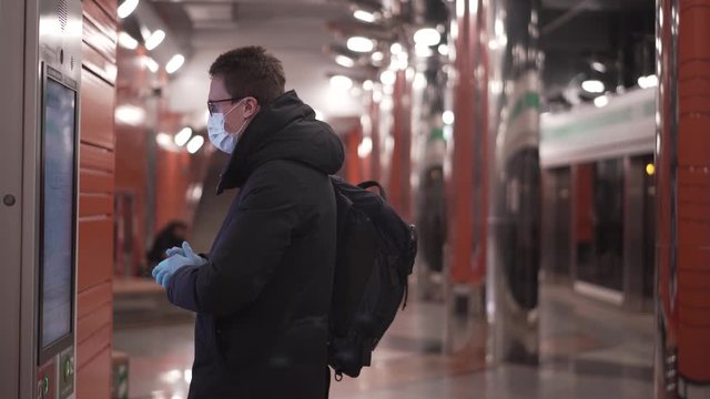 Young man standing in subway buying train tickets from a terminal wearing a face mask and medical gloves during the quarantine pandemic period