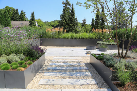 Moderne Garten- und Terrassengestaltung im Materialmix:Terrassen aus Steinplatten und Schotter sowie Metall Pflanzgef&auml;&szlig;e mit Gr&auml;sern, B&auml;umen und bl&uuml;henden K&uuml;bel Pflanzen	