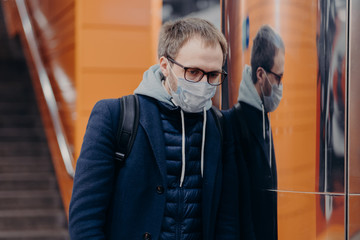 Sad infected man wears protective medical mask against coronavirus, poses in metro or subway, protects himself from infectious disease, carries backpack. Public transport during Covid-19 in Europe