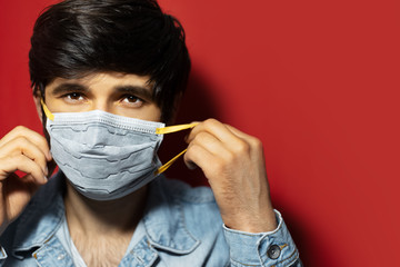 Studio portrait of young sick guy in denim jacket, put on medical face mask against coronavirus, isolated on red background.