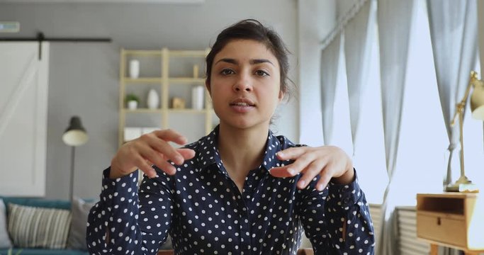 Head shot focused young indian woman sitting at desk, looking at camera, communicating via video call with client. Concentrated millennial female professional tutor recording giving online class.