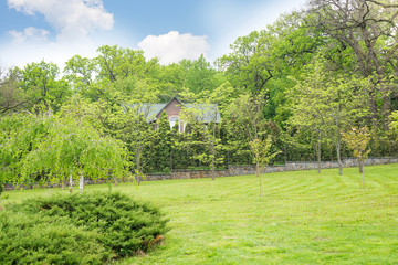 Park landscape with fresh green lawn, big house among the trees, bushes and blue cloudy sky.