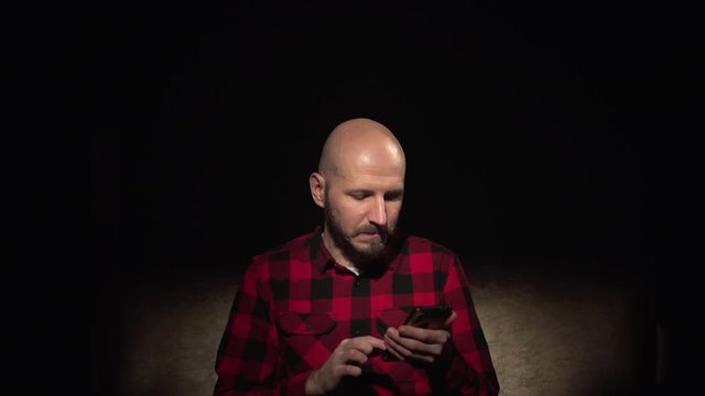 Young white bald hipster man with beard in plaid shirt sit on sofa read social media news on smartphone browsing isolated