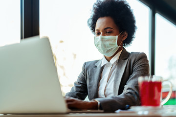 Black businesswoman typing on laptop while wearing face mask in a cafe.