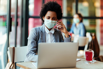 Black businesswoman with face mask talking on cell phone while using laptop in a cafe.