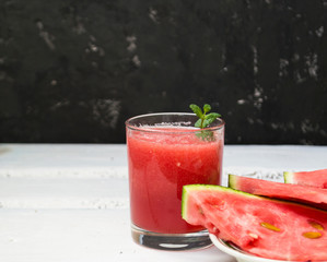 Slices of ripe watermelon on a plate and red watermelon juice in a glass on a wooden white background, and a black wall - side view. There is a place for text.