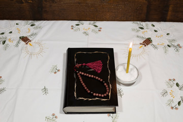 On the table is the book "Bible" in black binding. Above are the rosary. Nearby is a burning candle.