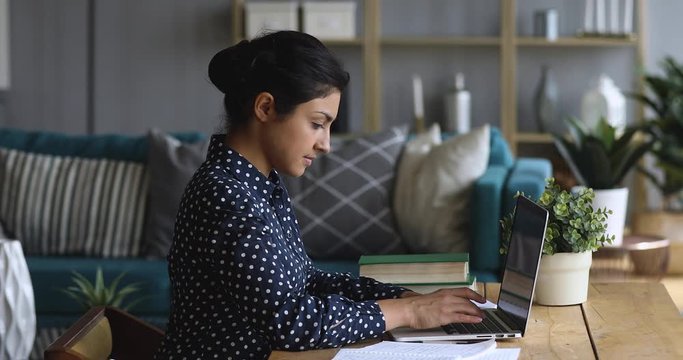 Side view concentrated millennial indian female college student woman doing university assignment or homework alone at home, using academic paper notes, doing educational project, sitting at table.