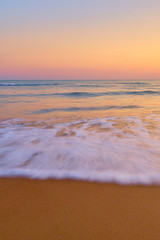 Colorful sunset at the tropical beach, sun behind clouds reflects on water and waves with foam hitting sand.