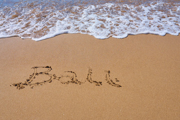 Bali inscription on a tropical sandy beach with waves and foam on a background.