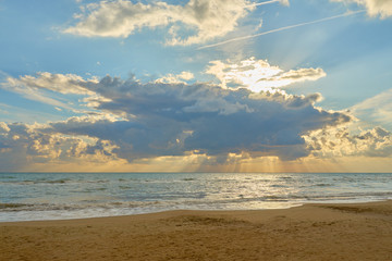 Colorful sunset at the tropical sandy beach, sun behind the clouds and waves with foam hitting sand.
