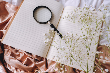 Spring composition, white gypsophila flowers with notebook and magnifying glass on the gold satin fabric