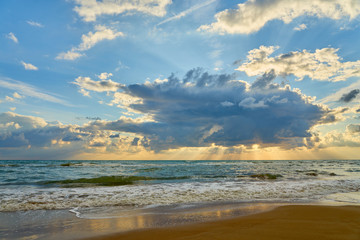 Colorful sunset at the tropical sandy beach, sun behind the clouds and waves with foam hitting sand.
