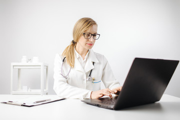 Concentrated mature woman in medical uniform and eyeglasses working with laptop while sitting at own office. Female doctor with stethoscope on neck using computer at workplace.