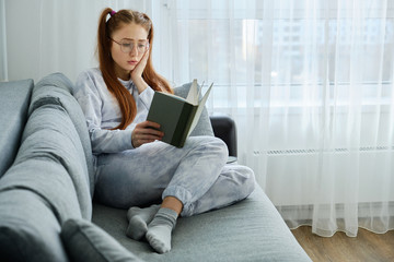 A red-haired girl with glasses, with long tails in pajamas sits on the couch and looks upset at the book, pressing palm to face