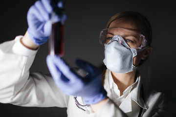 Concentrated female doctor in protective mask, eyewear and gloves holding glass bottle with blood...