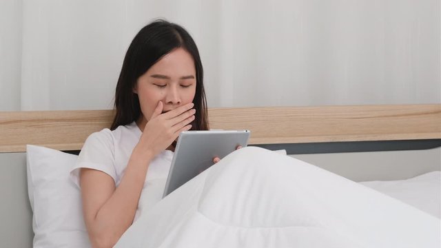 Beautiful Asian woman use tablet on bed and look sleepy by yawning then lie on white bed.
