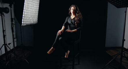 Sexy young model in stylish classic wear sitting on high bar stool and posing in dark photostudio