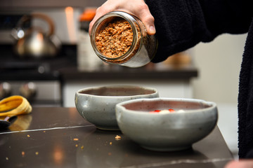 Pouring granola from a jar into a ceramic bowl while making a granola smoothie