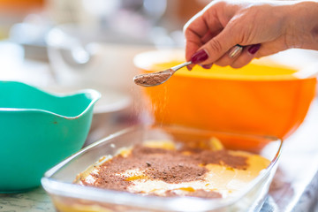 preparation of a tiramisu. Close up of a hand putting cocoa over the tiramisu cream with a teaspoon. Background is a kitchen table with bowls and bowls