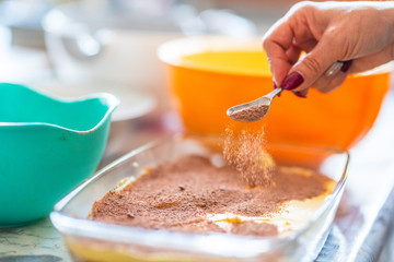 preparation of a tiramisu. Close up of a hand putting cocoa over the tiramisu cream with a teaspoon. Background is a kitchen table with bowls and bowls