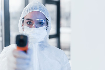 Holds infrared thermometer. Portrait of female doctor scientist in lab coat, defensive eyewear and...