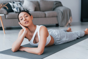 Fototapeta na wymiar Lying down on the mat and taking a break. Young woman with slim body shape in sportswear have fitness day indoors at home