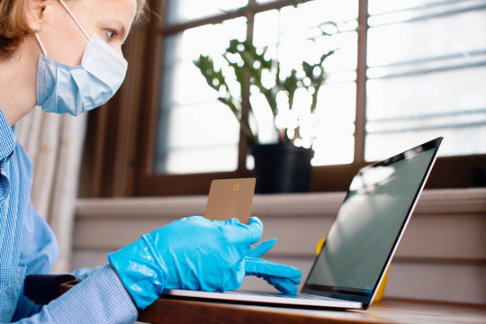 Person With Virus Protection Mask  And Gloves Holding Credit Card And Buying Online On Laptop Computer At Home By The Window