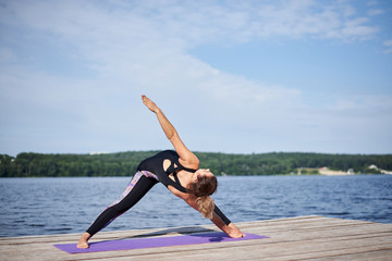 Young brunette woman with bare feet, wearing black and purple fitness outfit, stretching on violet yoga mat outside on wooden pier in summer. Fit girl, doing yoga poses by lake. Healthy lifestyle.