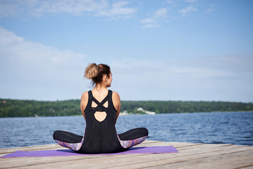 Fototapeta na wymiar Young brunette woman, wearing black and purple fitness outfit, sitting on violet yoga mat outside on wooden pier in summer. Fit girl, doing yoga poses by lake, thinking, relaxing, meditating