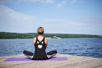 Fototapeta na wymiar Young brunette woman, wearing black and purple fitness outfit, sitting on violet yoga mat outside on wooden pier in summer. Fit girl, doing yoga poses by lake, thinking, relaxing, meditating