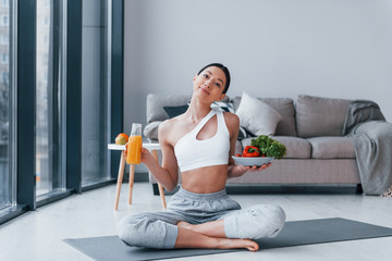 With healthy food and orange juice in hands. Young woman with slim body shape in sportswear have fitness day indoors at home