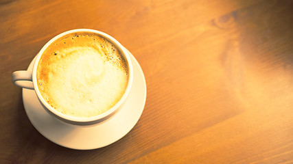 Cappuccino in white ceramic mug and plate on brown rustic wooden table. Cup of latte macchiato, black coffee with milk. Morning  beverage and drink. Coffee shop, cafe, bar atmosphere. Soft focus