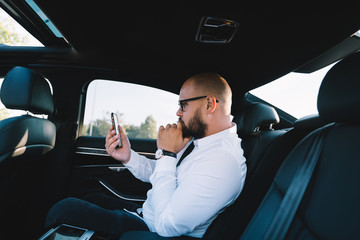 Side view of Caucasian male broker checking information on trade website connected to 4g wireless during business trip in rent auto car, formally dressed man texting during mobile communication