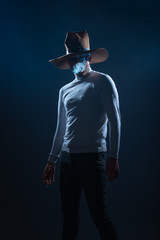 Portrait of a handsome young man with cowboy hat smoking cigarette in a studio on high contrast and...