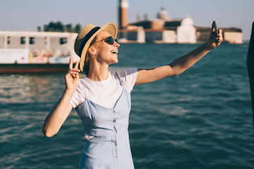 Happy female tourist in stylish sunglasses and straw hat clicking positive cellphone pictures for sharing media content to social networks and attract followers, cheerful traveller shooting web vlog