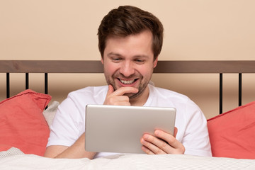 Young handsome man lying in bed and using tablet computer or reading an ebook