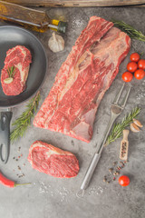 A large piece of beef cut into steaks, one of which is already in a pan with seasonings. Raw New York steak, top view.