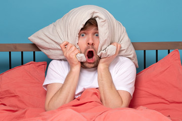 Cauacsian young man with pillow on head lying in bed and screaming