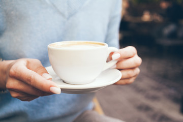 Close up of woman hands with beautiful white manicure holding cup of speciality coffee in restaurant terrace. Relaxing and boosting the mood. Lifestyle photo. Selective focus
