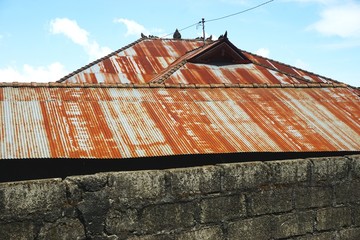 Rusty metal roof with dirty stone wall in foreground and blue sky behind in rural Indonesia