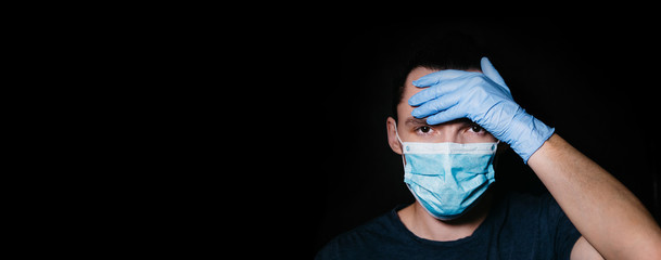 Coronavirus pandemic. Banner. A young man in a medical mask holds his forehead with his hand. Check body temperature. COVID - 2019. Virus protection. Latex gloves. Tired doctor in Italy