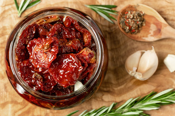 Sun-dried tomatoes with garlic, rosemary and spices in a glass jar on an olive wood cutting board, top view, flat lay