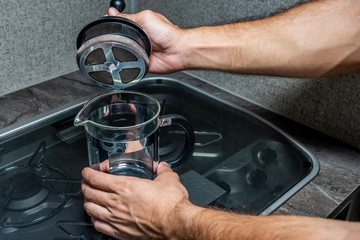Close up over the shoulder angle of a person's hands fixing coffee in a black and stainless steal metal french press over a small stove covered in glass and grey counter top inside one early morning.
