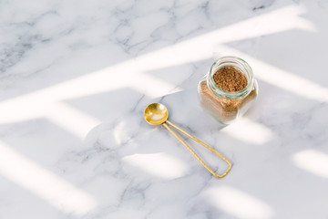 Facial powder ingredient in glass jar and brass spoon on white marble surface and dappled sunlight / Holistic wellness concept