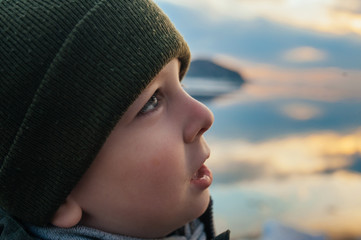 cute light-skinned boy in close-up with long black lashes, blue sparkling eyes and a knitted green hat looks rapturously at a beautiful blue-orange sunset on the sea.