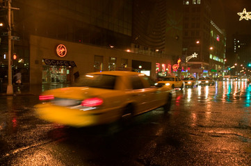 Speeding yellow taxi drives down rainy wet New York road at night with lights, New York
