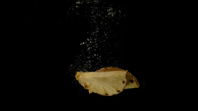 Two juicy slices of pineapple falling together in the dark clear water. Underwater steady slow motion camera shot.